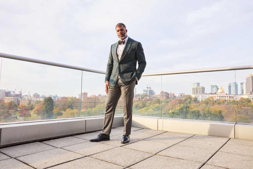 Tall handsome Black man is wearing a tuxedo and standing on a roof deck. You can see the Boston city skyline in the background.